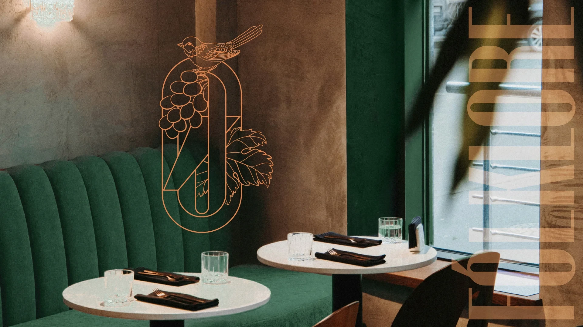 Intérieur du boutique-hôtel FÓLKLORE à Sancerre : banquette en velours vert, tables dressées et monogramme doré (oiseau et grappe de raisin)